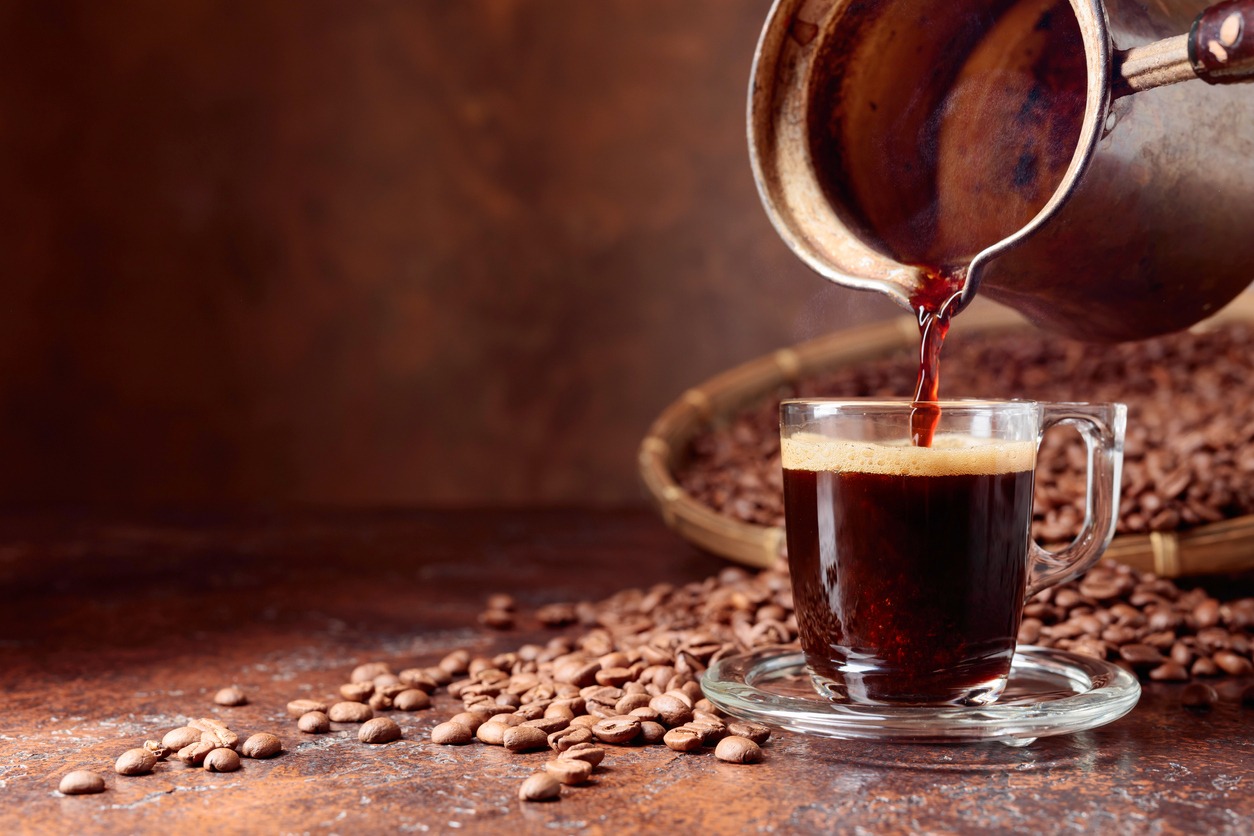 Black coffee is poured into a small glass cup from a old copper coffee maker.